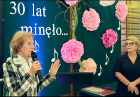 Pokaż zdjęcie: W tle napis 30 lat minęło, pry mikrofonie starsza kobieta.
