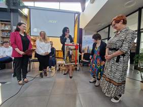 Pokaż zdjęcie: Goście i organizatorzy stoją w kawiarni. Elegancko ubrana kobieta przekazuje zdjęcie na którym jest osoba z zespołem Downa.