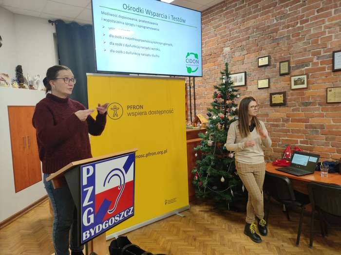 Na środku sali stoją: po lewej tłumaczka migowa, ubrana na czarno, a po prawej prelegentka - pracowniczka Kujawsko-Pomorskiego Oddziału PFRON. Widać także logo PZG, baner PFRON dot. dostępności oraz na ekranie - slajd z prezentacji dot. ośrodków wsparcia i testów