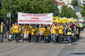 Pokaż zdjęcie: Osoby ubrane w żółte koszulki idą środkiem ulicy, w dłoniach trzymają instrumenty muzyczne bębenki i grzechotki, przed sobą niosą banery pierwszy z nich z napisem Marsz Godności Osób Niepełnosprawnych.