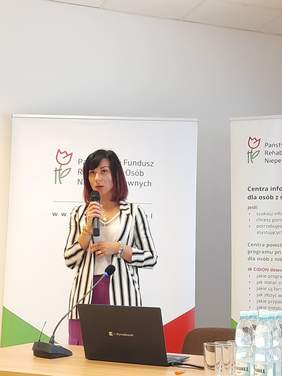 Pokaż zdjęcie: Kobieta przy mikrofonie ubrana w białą marynarkę w czarne paski, prowadzi wykład. W tle rollupy z logo PFRON.