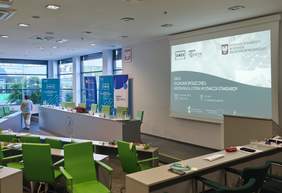 Pusta sala konferencyjna na Wydziale Ekonomii Uniwersytetu Szczecińskiego przed rozpoczęciem Gali Ekonomii Społecznej. Na pierwszym planie i po lewej widać rzędy biurek i jaskrawozielonych krzeseł. W głębi stoi stół prezydialny z godłem uczelni, a za nim trzy roll-upy organizatorów (w tym OWES i Fundacji Europa Lokalna). Nad stołem wisi duży ekran projekcyjny z wyświetlonym slajdem tytułowym: „GALA EKONOMII SPOŁECZNEJ: WSPÓŁPRACA, KTÓRA WYZNACZA STANDARDY”. Na bocznej ścianie widać galerię portretów. Pojedyncza osoba w białej bluzce przechodzi obok stołu prezydialnego.