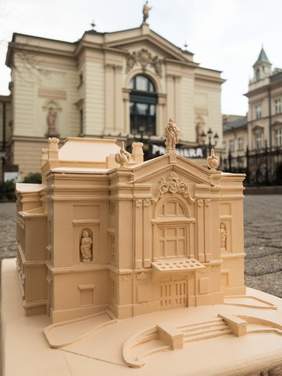 Pokaż zdjęcie: Na pierwszym planie makieta budynku Teatru Polskiego zrobiona dla osób niewidomych w tle widać niewyraźny zarys oryginalnego budynku teatru.