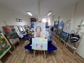 Przestronna sala. Wzdłuż ścian rozmieszczono wiele obrazów na sztalugach, krzesłach.