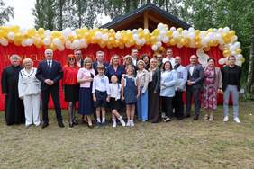 Pokaż zdjęcie: W plenerze stoi grupa osób. Są wśród nich dorośli i dzieci. Wszyscy są elegancko ubrani. Nad głowami ludzi są piękne girlandy z balonów. 