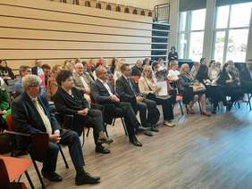 Pokaż zdjęcie: Na krzesłach ustawionych w półkole siedzą ludzie. Wszyscy patrzą przed siebie i słuchają wykładu. Są elegancko ubrani. 