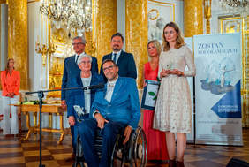 Pokaż zdjęcie: W Zamku Królewskim do pamiatkowego zdjęcia pozuje mężczyzna na wózku, laureat super lodołamacza, po jego lewej stronie stoi żona ubrana w jasna sukienkę, a po prawej stronie mama. Z tyłu dwaj mężczyźni w garniturach oraz kobieta w pomarańczowej sukience.