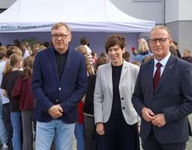 Oficjalne wydarzenie plenerowe. Od lewej stoją: Marek Machiński - dyrektor Szkoły Podstawowej nr 8 w granatowej marynarce, Anna Skupień - dyrektor Oddziału Wielkopolskiego PFRON w szarym żakiecie oraz prezydent Miasta Gniezna Michał Powałowski w garniturze z bordowym krawatem. W tle grupa młodzieży, biały namiot i ściana budynku.