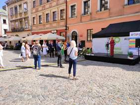 Pokaż zdjęcie: Na Rynku Starego Miasta w Lublinie, przed sceną zgromadzili się zaproszeni goście, w większości kobiety, trzymające w ręku torby prezentowe. Na scenie kobieta w czerwonym garniturze w kratkę, mówi do mikrofonu. Z tyłu za sceną stoją rollupy reklamowe z logo Regionalnego Ośrodka Polityki Społecznej.