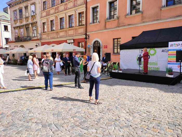 Pokaż zdjęcie: Na Rynku Starego Miasta w Lublinie, przed sceną zgromadzili się zaproszeni goście, w większości kobiety, trzymające w ręku torby prezentowe. Na scenie kobieta w czerwonym garniturze w kratkę, mówi do mikrofonu. Z tyłu za sceną stoją rollupy reklamowe z logo Regionalnego Ośrodka Polityki Społecznej.