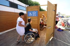 Pokaż zdjęcie: Na tarasie stoi przebieralnia wykonana z drewna z szerokimi drzwiami wjazdowymi oznaczonymi symbolem osoby poruszającej się na wózku w kolorze białym na niebieski tle. W przebieralni znajduje się niepełnosprawna osoba na wózku z opiekunem. Przed dr