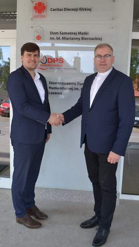 Pokaż zdjęcie: Dwóch mężczyzn podaje sobie dłoń. Stoją na tle budynku