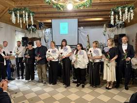 Pokaż zdjęcie: Grupa osób z różami stoi w udekorowanej sali z drewnianym sufitem, kwiatami i projektorem w tle, podczas uroczystości lub wydarzenia okolicznościowego.