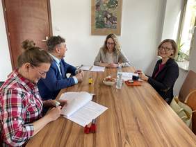 Pokaż zdjęcie: Przy długim drewnianym stole wraz z Prezes Fundacji siedzą mężczyzna ubrany w marynarkę i dwie kobiety. Wszyscy się uśmiechają. Trzymają w rękach dokumenty.