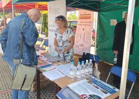 Pokaż zdjęcie: na zdjęciu widać stoisko z ulotkami i informacjami o dofinansowaniach ze środków PFRON na stoisku pracownik udziela informacji osobie która jest zaiteresowana pracą