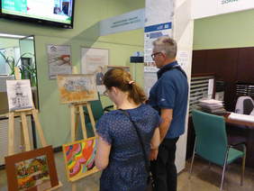 Pokaż zdjęcie: Na zdjęciu dwie osoby oglądają prace malarskie postawione na sztalugach, przedstawiające grafiki architektury oraz kwiaty i serca z nutami.