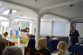 Pokaż zdjęcie: Sala z kolumnami. Z lewej strony są krzesła, w środku stół konferencyjny, z prawej strony mężczyzna w garniturze przemawia trzymając mikrofon.