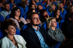 Widok na widownię kinową lub teatralną wypełnioną ludźmi. Na pierwszym planie skupia uwagę mężczyzna w okularach i ciemnej marynarce, patrzący z zaciekawieniem i powagą w stronę sceny. Obok niego, po lewej stronie, siedzi kobieta w białej kreacji z ciemnymi, upiętymi włosami, a po prawej kobieta w bluzce w czarno-białą kratkę. Wszyscy widzowie siedzą w niebieskich fotelach i są zwróceni w jednym kierunku.