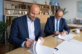 Pokaż zdjęcie: Dwaj mężczyźni w garniturach siedzą przy stole, uśmiechają się, lekko pochyleni, podpisują dokumenty