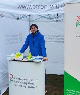 Pokaż zdjęcie: Stoisko informacyjne PFRON, a w nim pracownik w niebieskiej kurtce. Z przodu napis PFRON
