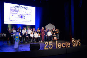 Pokaż zdjęcie: Zdjęcie przedstawia dużą grupę osób stojących i siedzących na wózkach inwalidzkich na scenie, podświetlonej niebieskim światłem. Z tyłu sceny znajduje się duży ekran z napisem "Jubileusz" i rysunkiem budynku, a poniżej niewyraźny tekst zawierający słowa "ŚRODOWISKOWEGO DOMU SAMOPOMOCY". Na pierwszym planie, w prawym dolnym rogu, widnieje duży, złoty napis: "25 lecie SDS". Osoby na scenie wydają się być uczestnikami lub organizatorami uroczystości jubileuszowej. Po lewej stronie sceny widoczna jest jedna kobieta przy pulpicie.