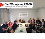Kilkanaście osób siedzi przy długim białym stole z rozłożonymi ulotkami. Na ścianie za uczestnikami wisi tablica z napisem „Państwowy Fundusz Rehabilitacji Osób Niepełnosprawnych”. W lewym górnym rogu widnieje hasło „Sieć Współpracy PFRON – działamy razem, by pomagać lepiej”. Po lewej stronie zdjęcia widać okno z pionowymi żaluzjami.
