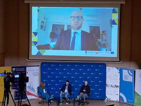 Pokaż zdjęcie: Zdjęcie zrobione z auli w kierunku panelistów. Jeden z panelistów jest online wyświetlany na ekranie, pozostałych troje siedzi w fotelach.