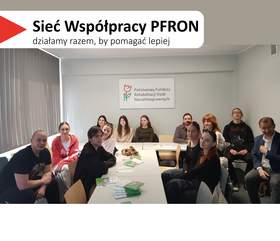 Kilkanaście osób siedzi przy długim białym stole z rozłożonymi ulotkami. Na ścianie za uczestnikami wisi tablica z napisem „Państwowy Fundusz Rehabilitacji Osób Niepełnosprawnych”. W lewym górnym rogu widnieje hasło „Sieć Współpracy PFRON – działamy razem, by pomagać lepiej”. Po lewej stronie zdjęcia widać okno z pionowymi żaluzjami.