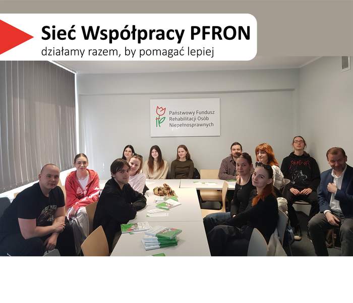 Pokaż zdjęcie: Kilkanaście osób siedzi przy długim białym stole z rozłożonymi ulotkami. Na ścianie za uczestnikami wisi tablica z napisem „Państwowy Fundusz Rehabilitacji Osób Niepełnosprawnych”. W lewym górnym rogu widnieje hasło „Sieć Współpracy PFRON – działamy razem, by pomagać lepiej”. Po lewej stronie zdjęcia widać okno z pionowymi żaluzjami.