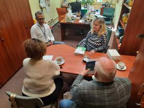 Pokaż zdjęcie: W gabinecie siedzą cztery osoby: dwie kobiety i dwóch mężczyzn. Rozmawiają ze sobą. Na stole leżą różne dokumenty, informatory, poradniki i filiżanki z herbatą. 