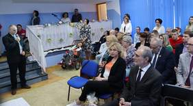 Pokaż zdjęcie: Zdjęcie osób uczestniczących w spotkaniu wielkanocnym, zaproszone osoby siedzą na krzesełkach, mężczyzna przemawia przez mikrofon. sala udekorowana jest świątecznie.   