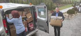 Kobieta z lewej części zdjęcia pakuje paczki do auta, obok przechodzi mężczyzna, który niesie paczkę 