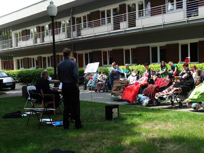 Pokaż zdjęcie: Na zdjęciu grupa osób, częśc siedzi przykryta kocem, kilka osób stoi, widac instrumenty muzyczne i dwóch artystów dających zebranym koncert