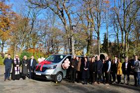 Pokaż zdjęcie: Grupa dwudziestu osób stoi w parku obok srebrnego busa przyozdobionego czerwoną kokardą oraz różowymi balonami. Przez korony drzew liściastych, z których opadły już liście, można dostrzec błękit bezchmurnego nieba.