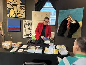 Kobieta i mężczyzna, kobieta ubrana w czarną bluzkę i czerwoną marynarkę, w ręce trzyma książkę w niebiesko-białej okładce, za stołem siedzi mężczyzna