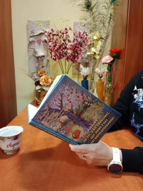 Pokaż zdjęcie: Kobieta trzyma w dłoniach podręcznik pt. "Jak żyć, a nie zwariować, gdy jedno z nas jest niepełnosprawne?". Na biurku stoi filiżanka. W tle widać wazoniki z kwiatami i inne ozdoby. 