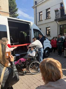 Pokaż zdjęcie: Kobieta na wózki inwalidzkim przecina czerwoną wstęgę na otwarcie nowo zakupionego busa do przewozu osób niepełnosprawnych.