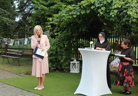 Pokaż zdjęcie: Kobieta w jasnoróżowym garniturze, trzymając mikrofon i teczkę z dokumentami, przemawia na tle zieleni podczas uroczystości jubileuszu WTZ w Siemiatyczach. Obok niej przy wysokim stole stoi siostra zakonna i uczestniczka warsztatu z upominkiem.