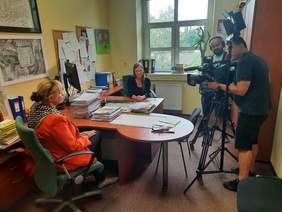 Pokaż zdjęcie: Przy stole siedzą naprzeciwko siebie dwie panie, które ze sobą rozmawiają. Po prawej stronie stoi dwóch mężczyzn, którzy nagrywają kamerami filmowymi kobiety. 