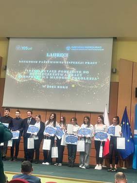 Pokaż zdjęcie: Grupa młodych osób pozuje na scenie, trzymając dyplomy i bony upominkowe. W tle widoczny jest ekran z napisem Laureaci konkursu Państwowej Inspekcji Pracy. Innowacyjne podejście do bezpieczeństwa pracy – perspektywa młodego pokolenia w 2024 roku.