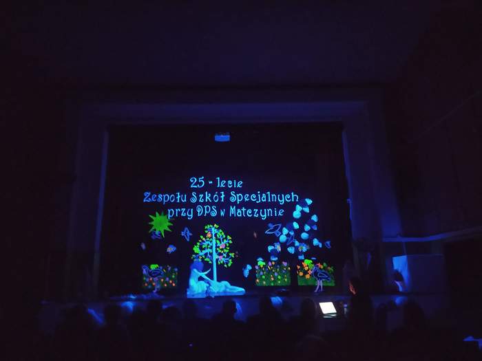 Pokaż zdjęcie: Zaciemniona scena podświetlona na niebiesko z białym napisem 25-lecie Zespołu Szkół Specjalnych przy DPS w Matczynie