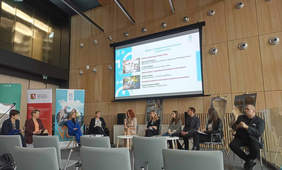 Pokaż zdjęcie: Debata w sali konferencyjnej w półokręgu ustawione są krzesła na których siedzą uczestnicy debaty, na ścianie wyświetlana jest prezentacja.