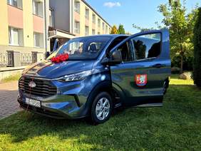 Pokaż zdjęcie: Na trawie przed ośrodkiem stoi niebieski bus z otwartymi drzwiami, udekorowany czerwoną kokardą.