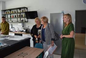 Pokaż zdjęcie: W pracowni kulinarnej, młody mężczyzna wałkuje ciasto, trzy kobiety oglądają pracownię i przyglądają się jego pracy