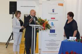 Pokaż zdjęcie: Mężczyzna z bukietem kwiatów stoi przed mównicą i przemawia do mikrofonu. Zanim stoi kobieta. Przed nimi stoi kobieta,która podpiera się kulą