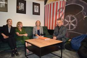 Pokaż zdjęcie: W pokoju relaksacyjnym na kanapach siedzą trzy kobiety i mężczyzna, na jednej ze ścian wiszą trzy czarno białe grafiki, na drugiej ścianie duże powiększenie taśmy filmowej