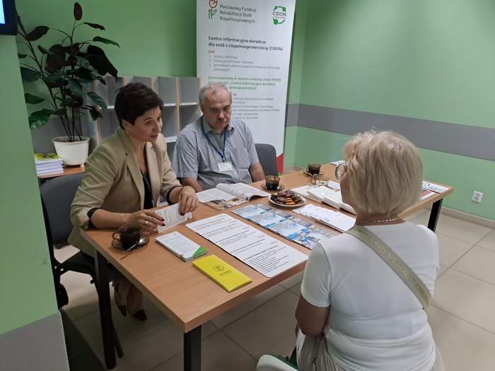 Pokaż zdjęcie: Przy biurku na którym leżą katalogi i ulotki siedzą trzy osoby, dwie kobiety i mężczyzna. Kobiety rozmawiają ze sobą. W tle baner z logo CIDON.