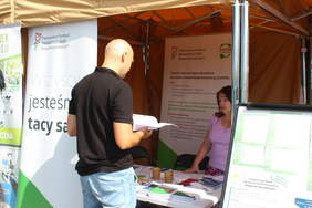 Pokaż zdjęcie: Przy stoisku informacyjnym stoi mężczyzna tyłem, kobieta udziela mu informacji