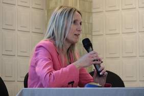Pokaż zdjęcie: Kobieta blondynka z długimi włosami i różowej marynarce, siedzi przy stole konferencyjnym, trzyma w ręku mikrofon, spogląda na salę konferencyjną
