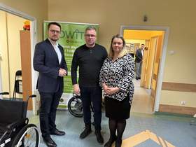 Na zdjęciu dwóch mężczyzn w ciemnych strojach i kobieta w biało czarnej bluzce z włosami w kolorze blond. po lewej stronie widoczny wózek inwalidzki, a w tle roll up z napisem OWIT